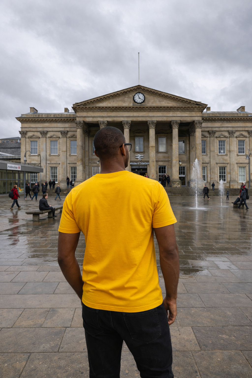 Person wearing a yellow shirt standing in front of Huddersfield Train Station, it's a large building with columns on a cloudy day.