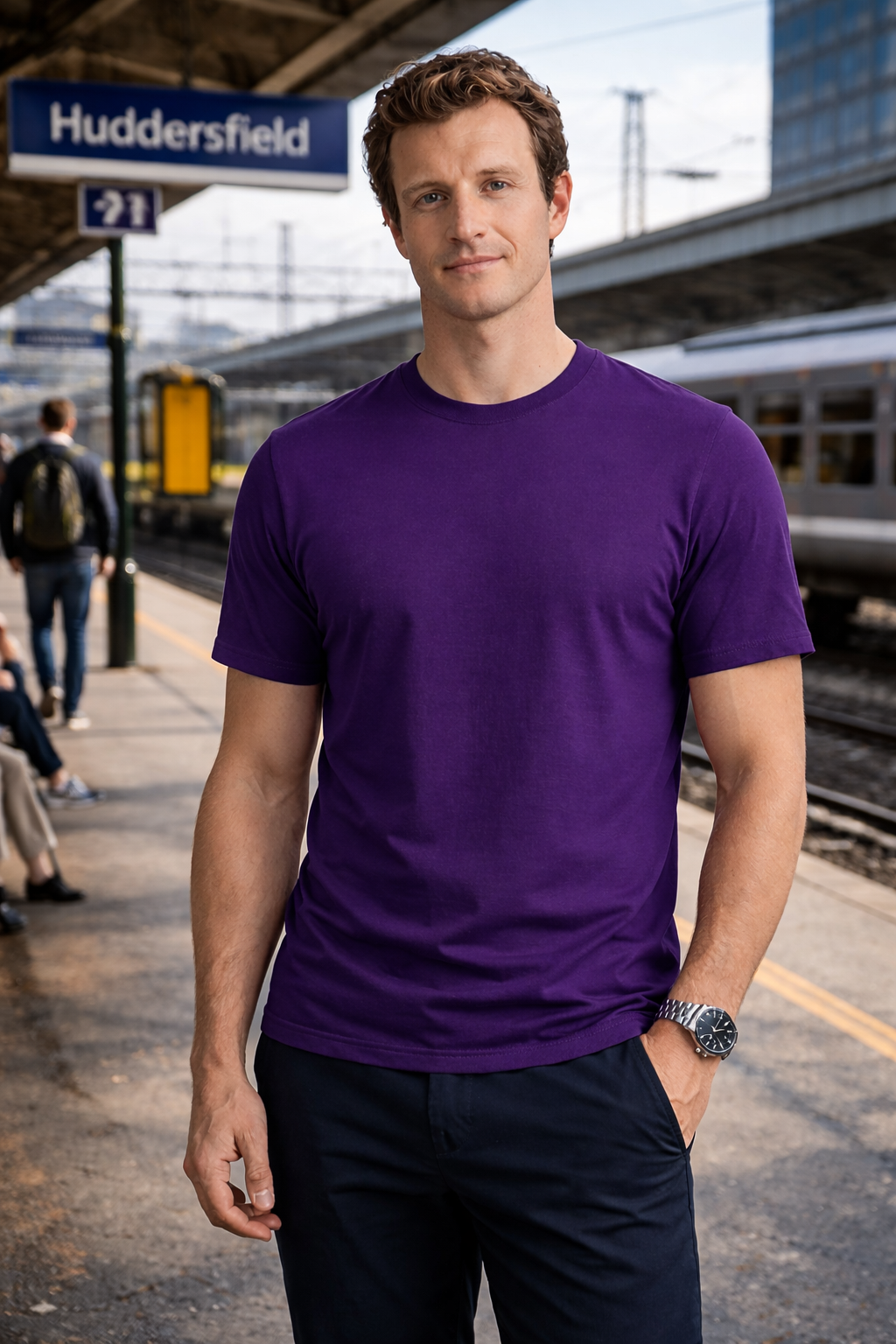 Man wearing a purple t-shirt at a train station with Huddersfield sign in the background.